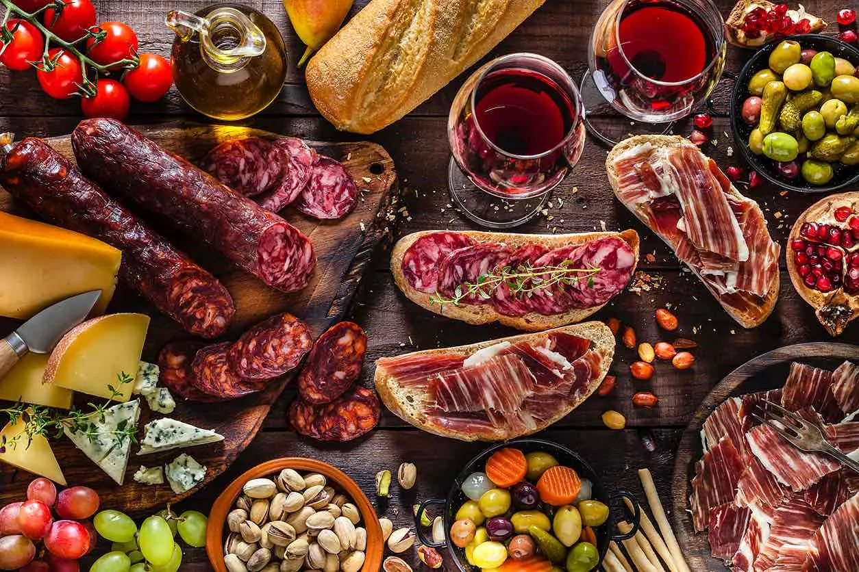 Koyu ahşap bir zeminde, iki kadeh kırmızı şarap eşliğinde, dilimlenmiş salam, jambon ve sosis gibi çeşitli şarküteri ürünleri, farklı peynirler, zeytin, taze üzümler, nar taneleri, kuruyemişler ve ekmek dilimleri içeren bol çeşitli ve iştah açıcı bir meze sofrası görülüyor.