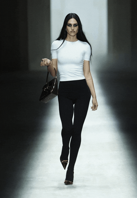 Gucci Sonbahar Kış 2026 defilesinde beyaz dar kesim tişört ve siyah tayt kombinasyonu, minimal çizgilerle güçlü bir siluet oluştururken; elde taşınan parlak siyah çanta ve sivri burun topuklular görünümü sofistike ve iddialı bir runway stiline dönüştürüyor.