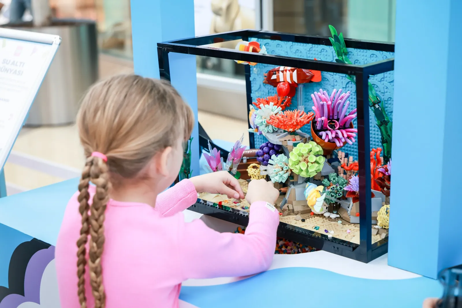 Bir kız çocuğunun pembe bluzuyla, arkası dönük bir şekilde renkli mercanlar, balıklar ve deniz bitkileriyle dolu bir su altı LEGO sergisinde yaratıcı bir şekilde parçaları düzenleyerek sanatsal üretim süreçlerine katılımını gösteren detaylı bir görüntü.