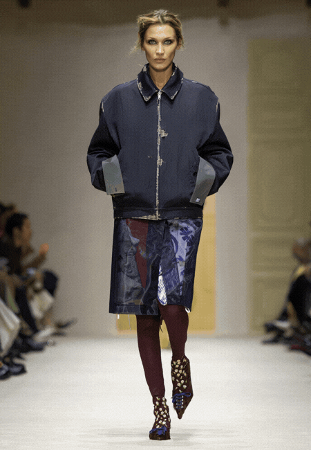 Prada Sonbahar Kış 2026 defilesinde lacivert oversize ceket, desenli diz altı etek ve bordo çorap kombinasyonu ile güçlü kontrast ve modern siluet sunan kadın runway görünümü.