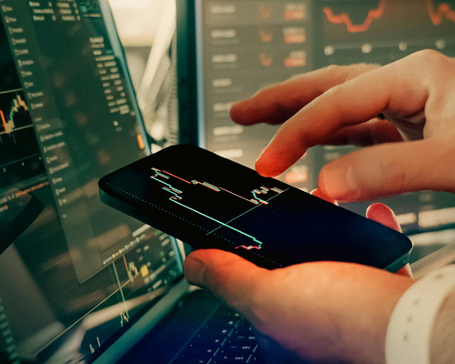 Bir kişinin eli, üzerinde finansal grafikler ve veriler gösteren bir akıllı telefonu tutuyor; arka plandaki çoklu bilgisayar ekranlarında da benzer borsa ve yatırım analizleri yer alırken parmak ekrana dokunarak dijital piyasaları yönetiyor.