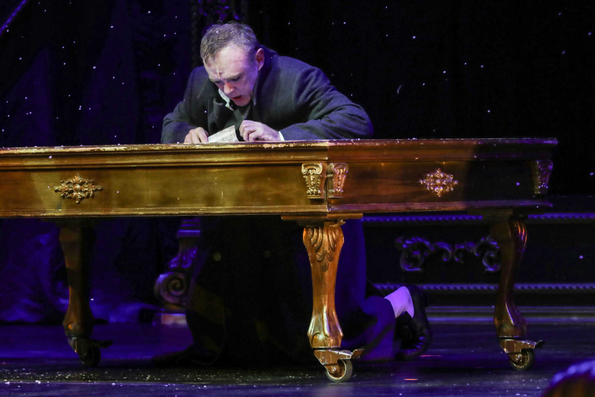 Sahnenin karanlık ve mor tonlarla aydınlatılmış atmosferinde, Amadeus oyununda Antonio Salieri'yi canlandıran oyuncu, dönem kostümüyle altın varaklı gösterişli bir masanın arkasında diz çökmüş, elindeki nota kağıtlarına yoğun ve ızdırap dolu bir ifadeyle bakarak Mozart'ın dehası karşısındaki içsel mücadelesini yansıtıyor.