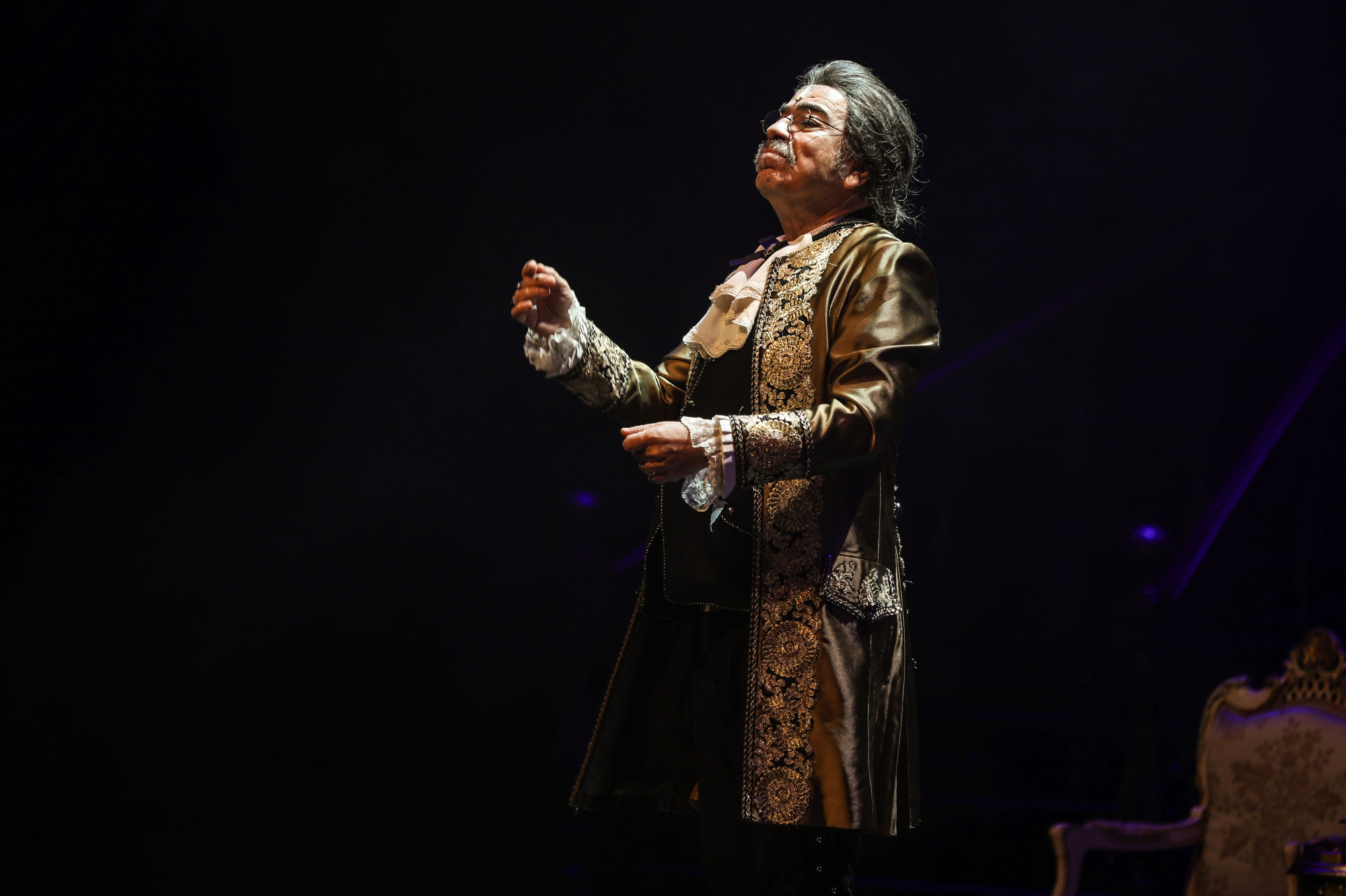 Sahne ışıkları altında, Amadeus oyununda Antonio Salieri karakterini canlandıran Selçuk Yöntem, altın işlemeli, 18. yüzyıl dönemi kostümüyle, başı hafifçe yukarıda ve elleri havada, deha ile kıskançlık arasındaki karmaşık duyguları yansıtan etkileyici bir performans sergiliyor.
