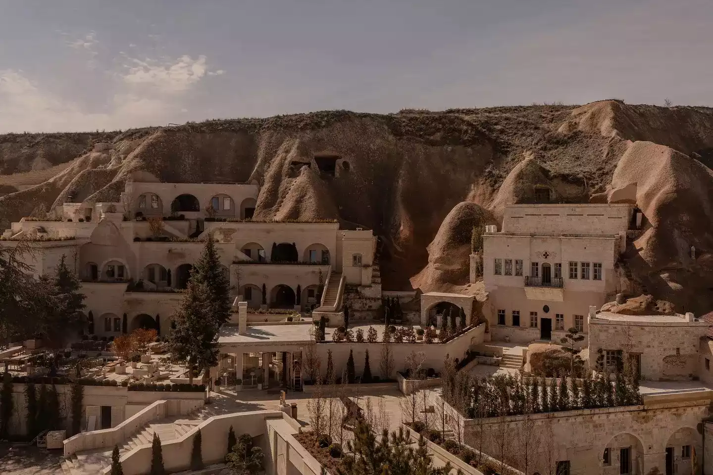 Doğal kaya oluşumlarına ustaca entegre edilmiş, avangart mimarisi ve zarif tasarımıyla dikkat çeken, çok katlı taş binalardan oluşan, teraslar ve kemerli pencerelerle süslü, Kapadokya'ya özgü lüks bir tesisin geniş bir görüntüsü.