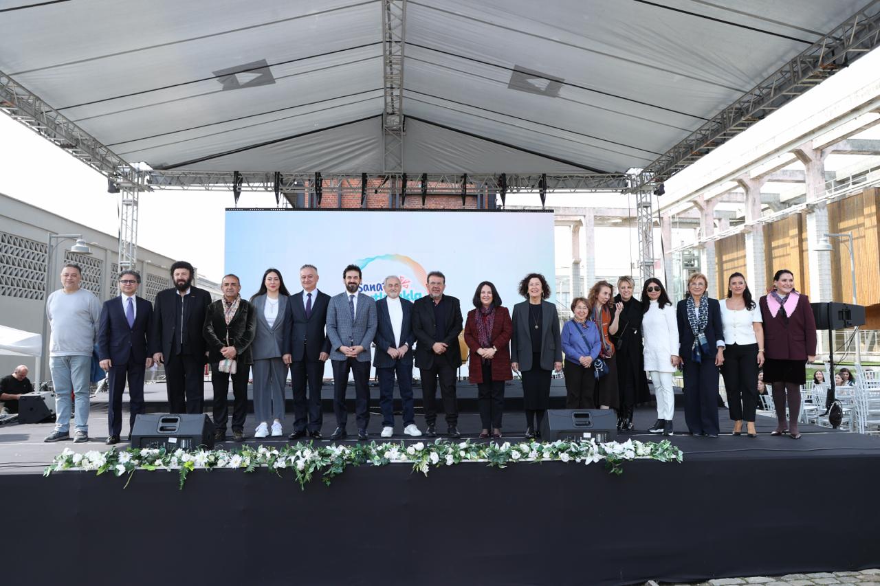 Beyaz bir çatı altında, önünde beyaz çiçek ve yeşillik süslemeler olan sahnede, arkalarında Sanatla Gelecek yazılı ekranın bulunduğu bir Bienal açılışında poz veren, takım elbiseli erkekler ve çeşitli kıyafetlerdeki kadınlardan oluşan kalabalık bir grup.