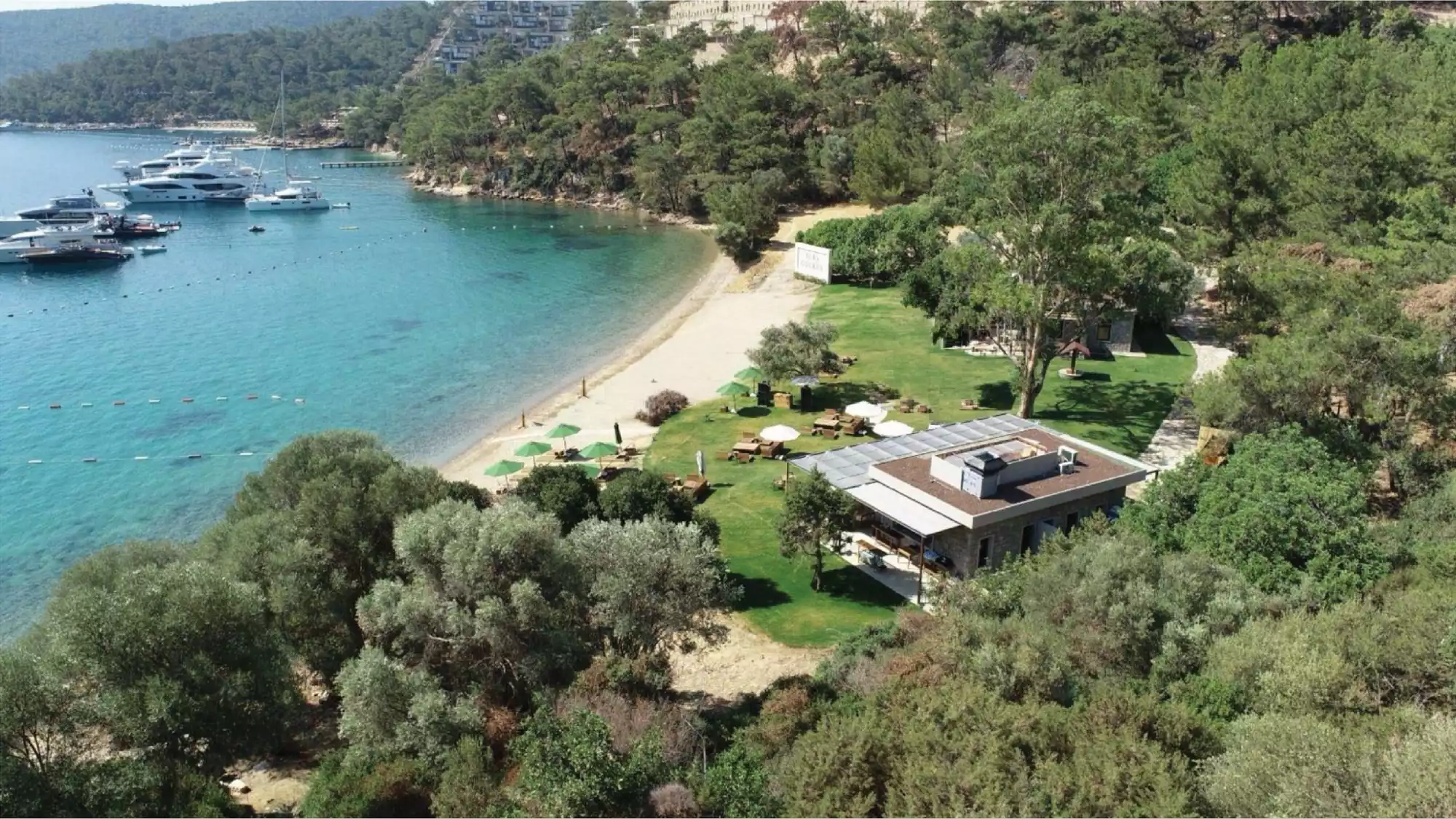 Lüks yatların demirli olduğu berrak turkuaz sulara sahip sakin bir koyun kıyısında, şemsiyeli kum plajı, yeşil çimenlik alanı, açık hava oturma yerleri ve modern bir yapının da bulunduğu, yoğun Akdeniz bitki örtüsüyle kaplı tepelerin göründüğü büyüleyici bir sahil tesisi manzarası.