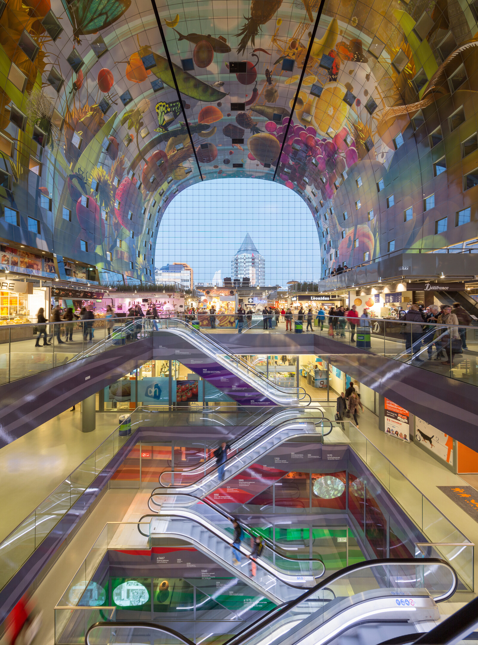Rotterdam'ın ikonik Markthal'ının içinden çekilen geniş açılı bu fotoğrafta, devasa meyve, sebze ve böcek motifleriyle süslü kavisli tavanın altında, katlar arasında hareket eden sayısız yürüyen merdiven ve canlı pazar dükkanları görülüyor.