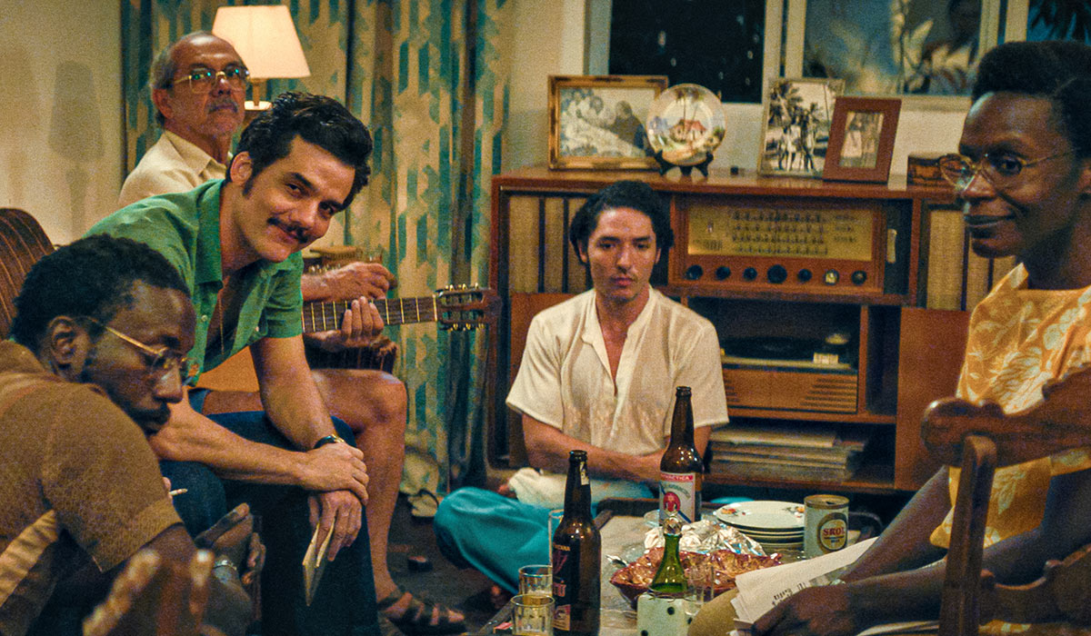 Wagner Moura, yeşil gömlekli ve bıyıklı haliyle, bir grup insanla samimi bir ev ortamında gitarıyla birlikte otururken doğrudan kameraya gülümseyerek bakıyor; etrafta içecekler, yiyecekler, eski bir radyo ve çerçeveli fotoğraflar bulunan bu sahne muhtemelen Wagner Moura'nın The Secret Agent filmindeki bir rolünü yansıtıyor.