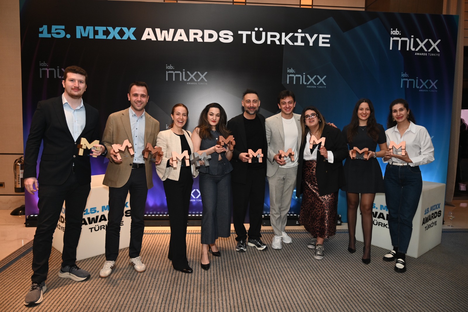 15. Mixx Awards Türkiye ödül töreninde, M harfi şeklindeki çeşitli renklerdeki ödüllerini elinde tutan dokuz kişilik bir ekip, etkinliğin logosunun yer aldığı şık bir sahne önünde neşeli ve gururlu bir şekilde gülümsüyor.