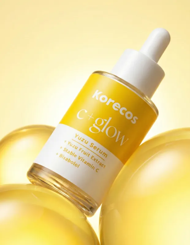 Parlak sarı bir arka planda, Korecos C+Glow Yuzu Serum yazılı, sarı etiketli damlalıklı şişe, cilde aydınlık bir görünüm vadeden Yuzu Meyve Özü ve Stabil C Vitamini içeren yenilikçi bir cilt bakımı gelişmesini temsil eden şeffaf sarı kürelerin üzerinde eğik duruyor.