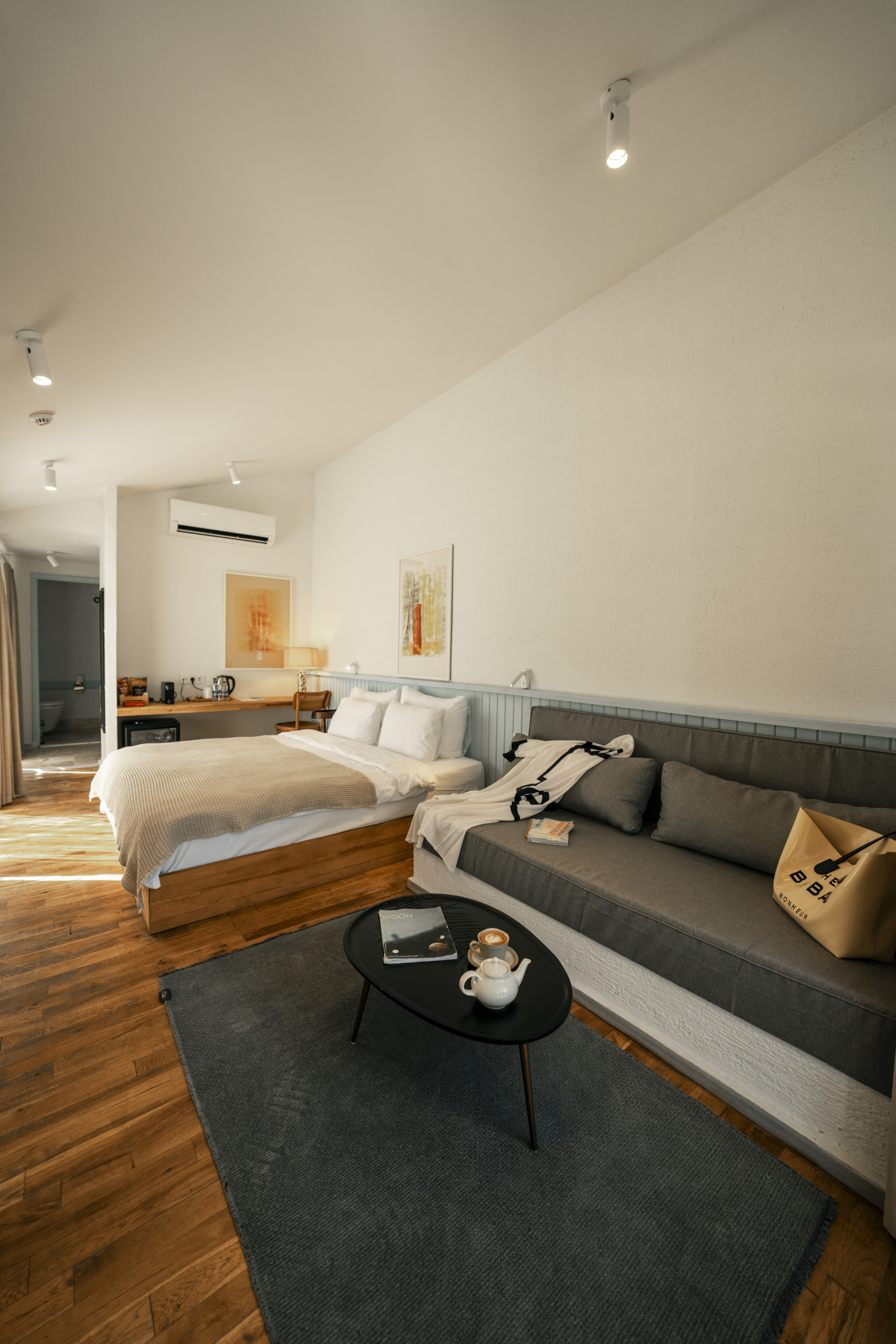 Nayu İğneada'daki modern bir otel odasının iç mekanı, büyük bir ahşap yatak, rahat bir gri kanepe, sehpa üzerinde dergi ve kahve fincanı ile İğneada'nın huzurlu atmosferini yansıtan aydınlık, ahşap zeminli bir konaklama birimini gösteriyor.
