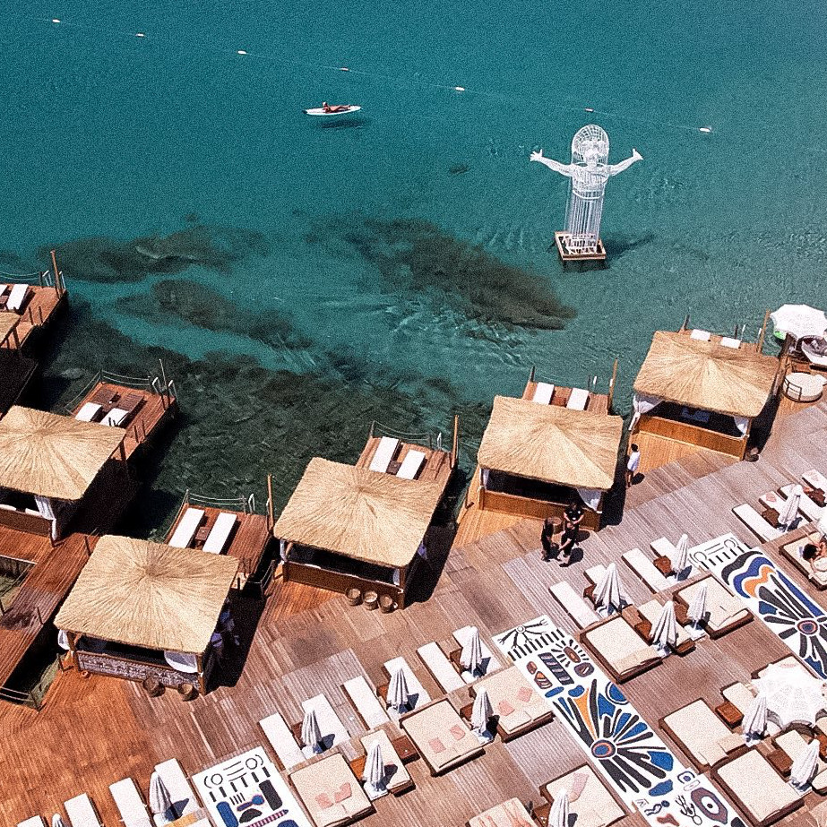 Bir beach club'ın kuşbakışı görüntüsü, berrak turkuaz denizde kolları açık beyaz bir heykelin yükseldiği, ahşap iskele üzerinde hasır çatılı localar ve şezlongların doğal ve şık bir tatil atmosferi sunduğu Çeşme'deki The Beach of MOMO Dalyan'ı gösteriyor.