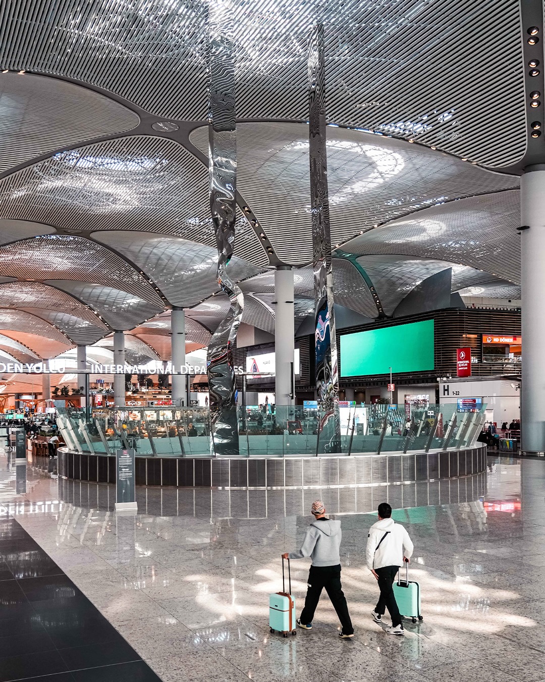 Modern ve geniş İstanbul Havalimanı terminalinde, dalgalı ve ışıltılı tavan yapısının altında, iki yolcu parlak mozaik zemin üzerinde tekerlekli, açık mavi valizleriyle Uluslararası Gidiş tabelalarına doğru ilerliyor.