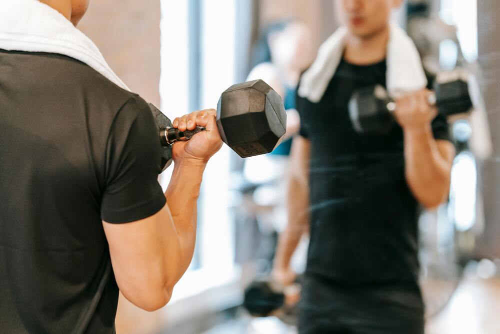 Bir aynanın karşısında, siyah bir tişört ve omzunda beyaz bir havluyla kol kaslarını çalıştıran bir adam, fitness rutininin ve yoğun bir spor aktivitesinin tam ortasında, spor sonrası beslenmenin önemini vurgulayan anı yansıtıyor.