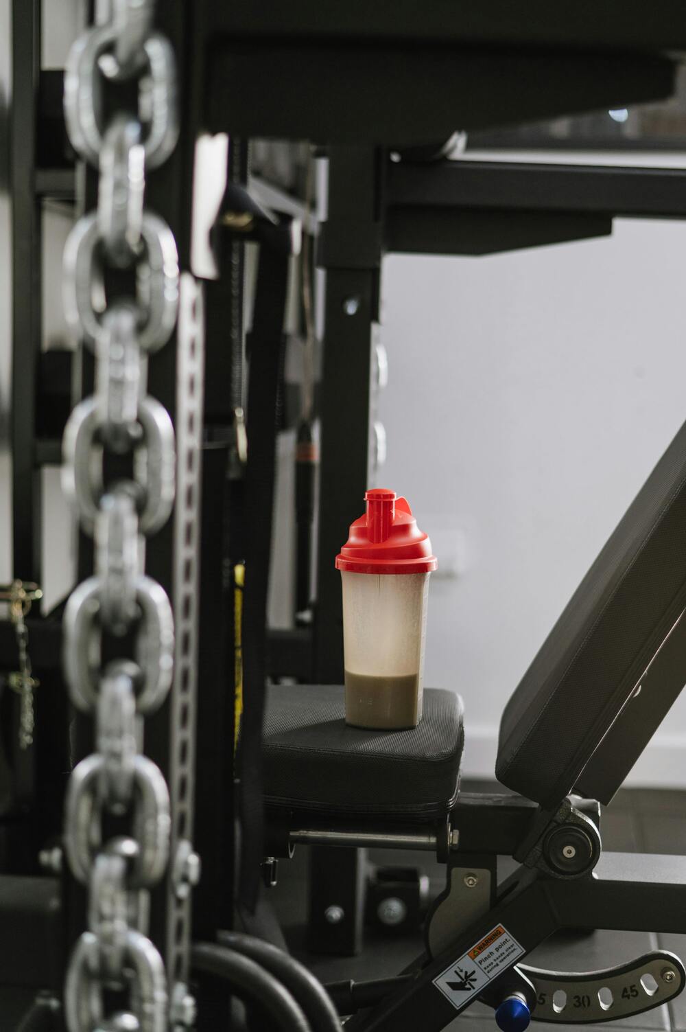 Siyah bir spor sehpası üzerinde duran kırmızı kapaklı, içinde kahverengi bir içecek bulunan şeffaf shaker şişesi, spor sonrası beslenme pratiğini gösteren bir spor salonu ortamında, bulanık zincirler ve ekipmanlarla çevrili.
