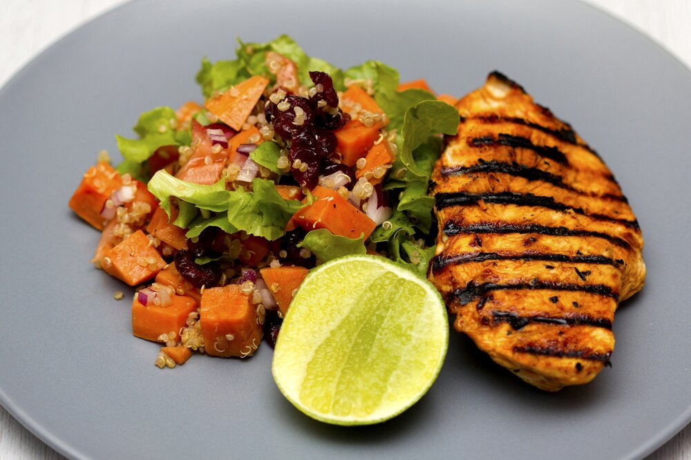 Gri bir tabakta, protein açısından zengin ızgara tavuk göğsü, enerji veren kinoa, tatlı patates, taze marul, yaban mersini ve kırmızı soğan içeren renkli bir salata ile yanında dilimlenmiş misket limonu, spor sonrası beslenme için ideal, dengeli ve sağlıklı bir öğün sunuyor.