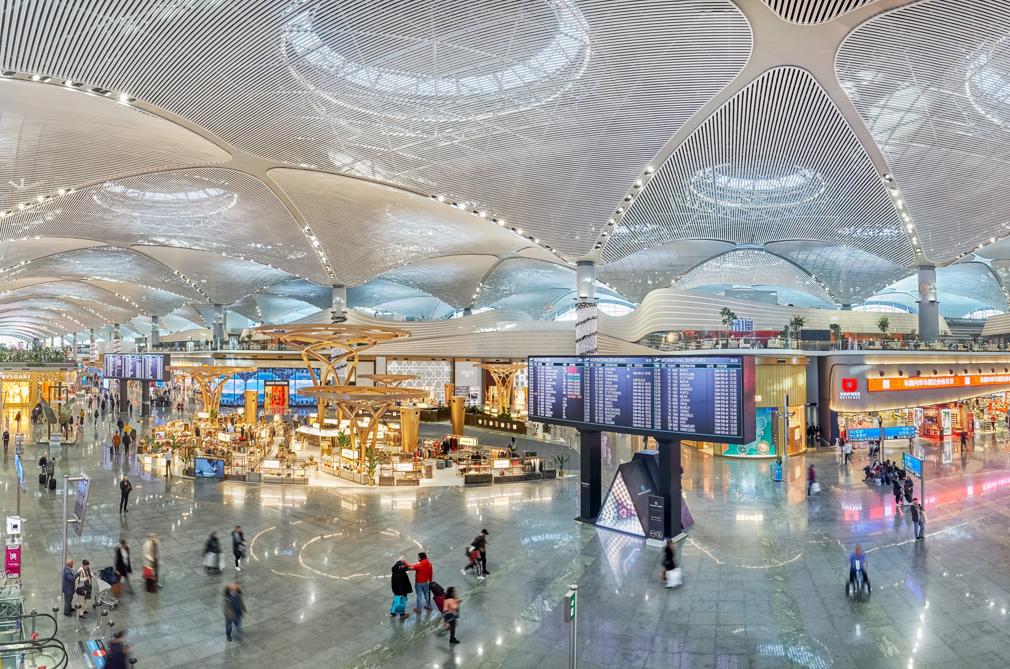 Modern mimariye sahip geniş ve hareketli bir havaalanı terminali içinde, yansıtıcı parlak zeminde dolaşan birçok yolcu, kendine özgü yüksek, dalgalı tavanın ve belirgin bir Unifree Duty Free mağazasının yanı sıra lüks dükkanlar ile dijital uçuş bilgi panolarının çevrelediği canlı alanı gösteriyor.