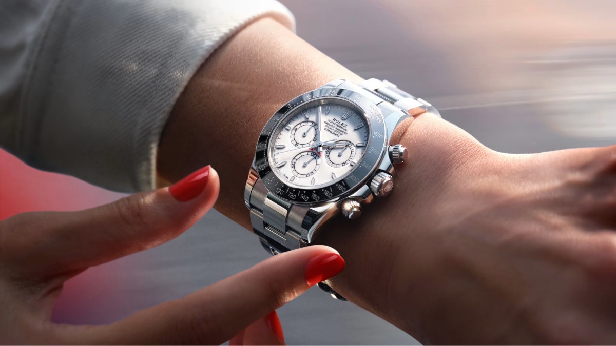 Kırmızı ojeli bir elin hafifçe uzandığı bir bilekte, parlak metal kordonlu ve beyaz kadranlı, ikonik Rolex Cosmograph Daytona kronograf saat tüm detaylarıyla göz alıyor, modern tasarımı ve fonksiyonel sub-kadranları dikkat çekiyor.