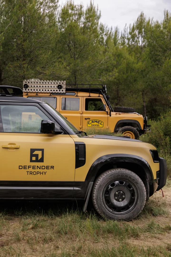 Ön planda çimenli arazide park etmiş sarı renkli, siyah detaylı yeni nesil bir Defender Trophy aracı, arka planda ise ağaçlar arasında park etmiş eski model Camel Trophy logolu sarı bir Defender off-road aracı görünüyor, bu görüntü geçmişle bugünü bir araya getirerek heyecan verici bir Defender Trophy macerasına işaret ediyor.
