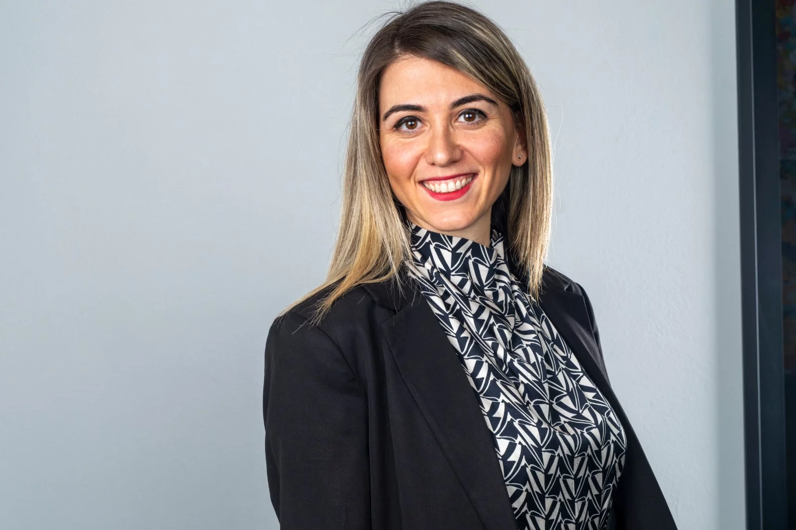 Parlak gülümsemesi, omuzlarına dökülen sarı ve kahve tonlarındaki saçları ve kırmızı rujuyla poz veren Beste Toksoy, şık siyah bir blazer ceketinin altında modern siyah beyaz desenli bir bluz giymiş, açık gri bir stüdyo arka planında doğrudan kameraya bakıyor.