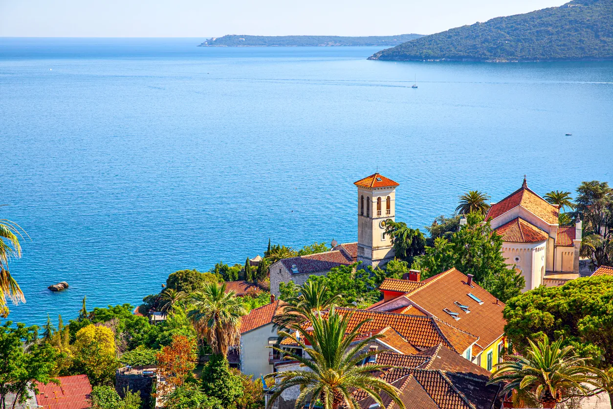 Adriyatik Denizi'nin berrak suları ve uzak adalarla çevrili Herceg Novi'de, güneşli bir günde, kırmızı kiremitli çatılara sahip geleneksel evlerin, yemyeşil palmiye ağaçlarının ve tarihi saat kulesinin panoramik manzarası, bu pitoresk kıyı şehrinin güzelliğini sergiliyor.