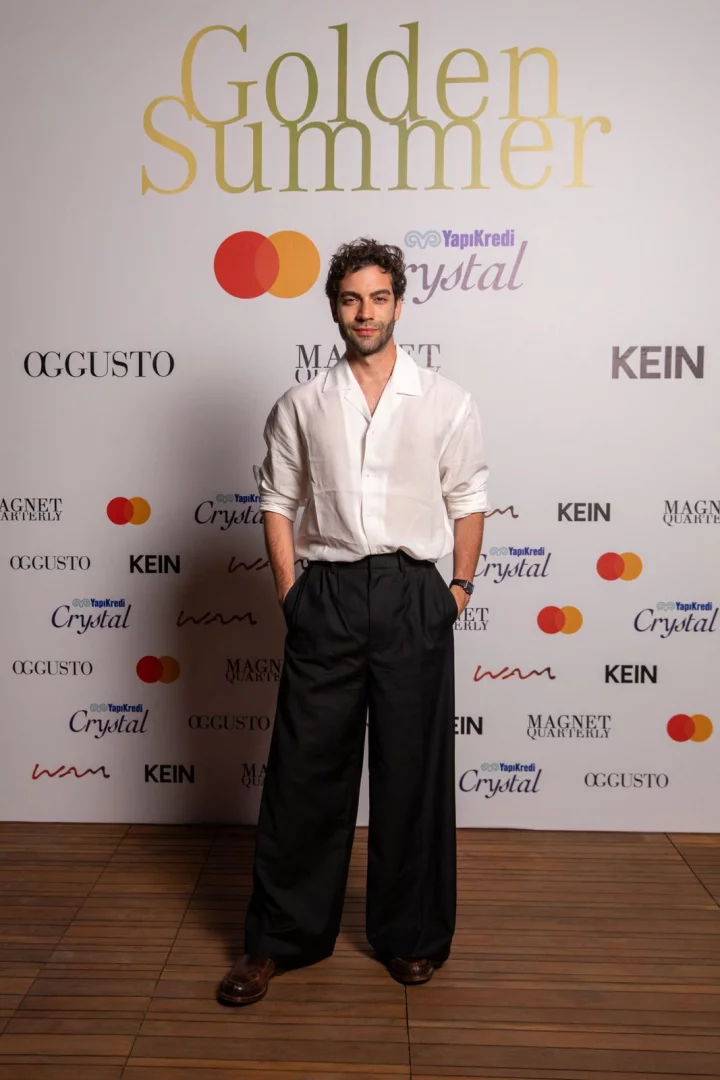 Beyaz Golden Summer yazılı ve çeşitli sponsor logolarıyla dolu bir fonda ayakta duran, beyaz bol bir gömlek, siyah geniş paça pantolon ve kahverengi ayakkabılar giyen, elleri ceplerinde, kameraya hafifçe gülümseyerek bakan kıvırcık saçlı genç bir adam.