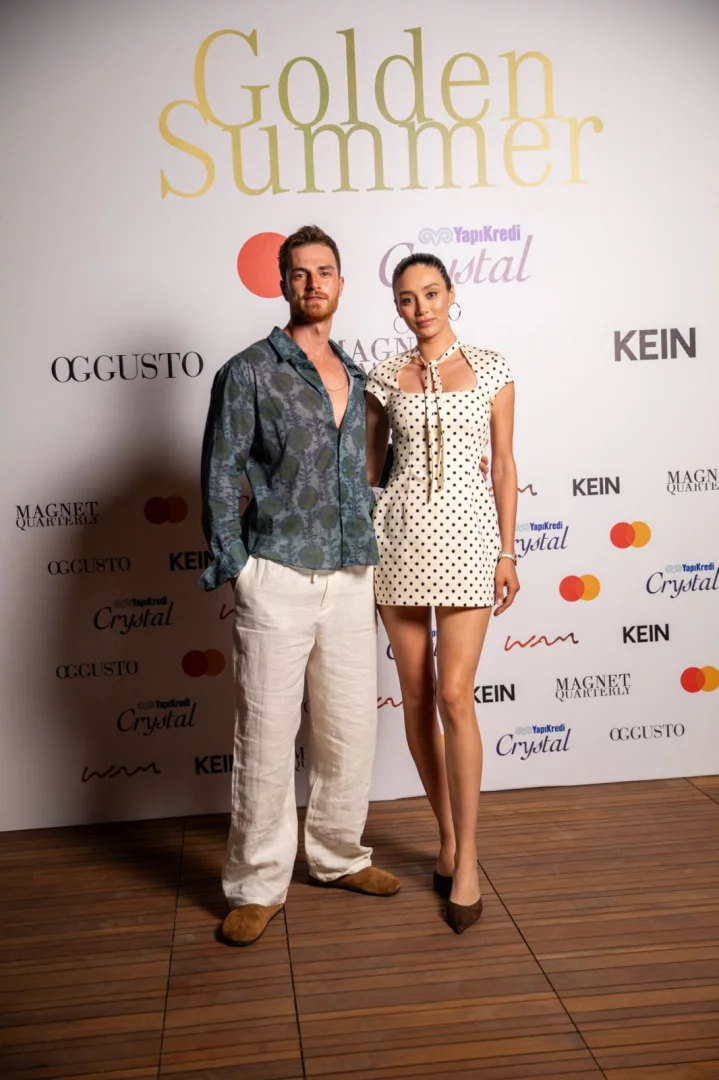 Golden Summer etkinliğinde, arkasında çeşitli sponsor logolarının bulunduğu beyaz bir fonun önünde, desenli yeşil gömlek ve krem rengi keten pantolon giymiş sakallı bir erkek ile boyun kısmında bağlama detayı olan puantiyeli beyaz mini elbise giymiş, saçları toplu bir kadının birlikte poz verdiği net bir görüntü.
