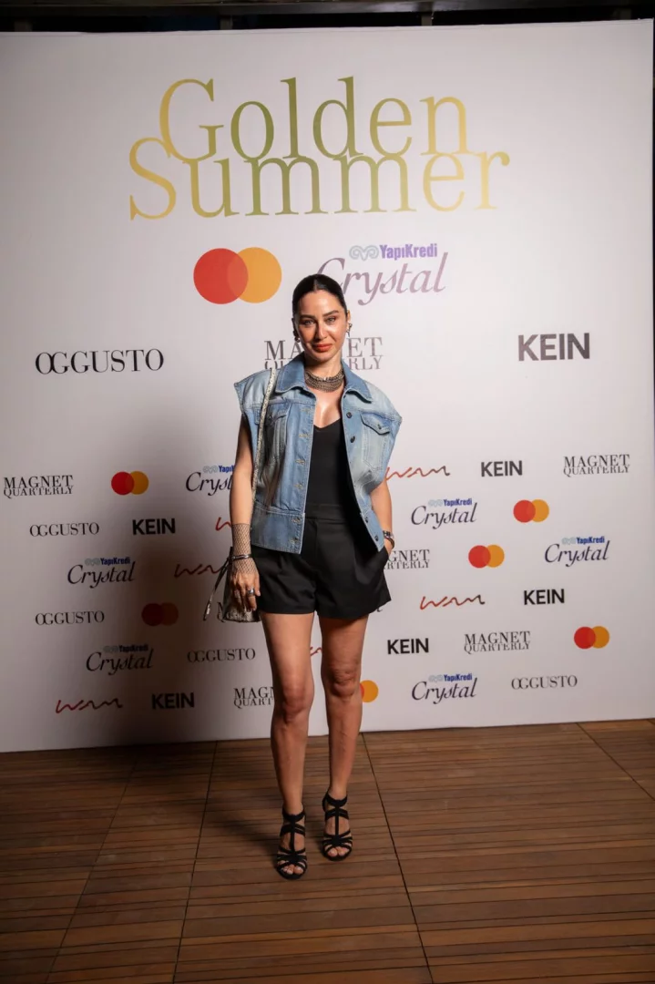 Altın rengi büyük harflerle yazılmış Golden Summer başlığı ve Yapı Kredi Crystal, Mastercard gibi sponsor logolarıyla kaplı beyaz bir fonun önünde, siyah şortu, kot yeleği ve bantlı topuklu ayakkabılarıyla modern ve şık bir görünüm sergileyen kadın, hafifçe gülümseyerek poz veriyor.
