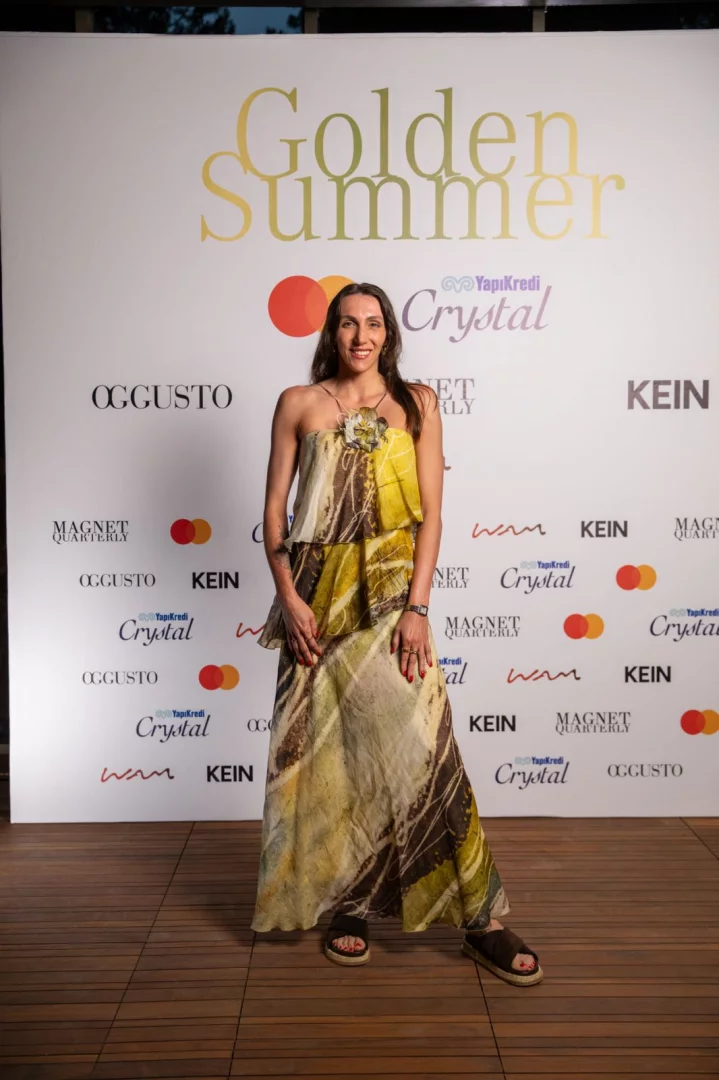 Bodrum Demirbükü’nde gerçekleşen Golden Summer davetinde, beyaz fon önünde desenli askısız bir elbise ve rahat sandaletlerle şık bir duruş sergileyen, uzun koyu saçlı bir kadın zarifçe gülümsüyor, arkasında Yapı Kredi Crystal, OGGUSTO ve Mastercard gibi iş birliği partnerlerinin logoları Akdeniz yaşam kültürünü yansıtan bu etkinliğin ruhunu vurguluyor.