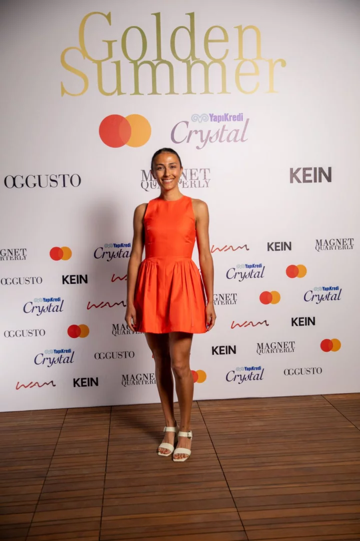 Gülümseyen, koyu saçları toplanmış, turuncu askılı kısa bir elbise ve beyaz topuklu sandaletler giyen bir kadın, Bodrum'da düzenlenen Golden Summer etkinliğinde, OGGUSTO, Magnet Quarterly, KEIN, Mastercard ve Yapı Kredi Crystal logolarının tekrarlandığı büyük altın renkli Golden Summer yazılı beyaz bir fotoğraf duvarının önünde duruyor.