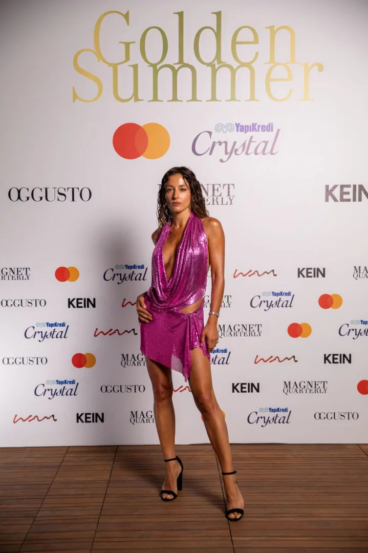 Dalgalı kahverengi saçlı, derin dekolteli ve asimetrik etekli parıltılı fuşya örgü mini elbise giymiş bir kadın, Golden Summer etkinliği markası ve YapıKredi Crystal gibi sponsor logolarının bulunduğu beyaz bir fon önünde kendinden emin bir şekilde poz veriyor.
