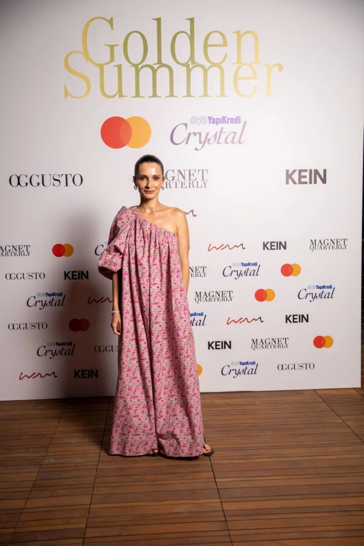 Golden Summer temalı bir etkinlikte, üzerinde çeşitli sponsor logolarının yer aldığı beyaz panonun önünde, pembe çiçek desenli, tek omuzlu kabarık kollu uzun bir elbise giyen zarif bir kadın ahşap zeminde durarak hafifçe gülümsüyor.
