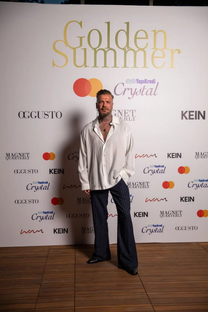 Boynunda ve göğsünde dövmeler olan, beyaz bol gömlek ve lacivert çizgili İspanyol paça pantolon giyen bir adam, sol eli cebinde, arkasında Golden Summer başlığı ve YapıKredi Crystal gibi sponsor logolarının tekrarlandığı beyaz bir panonun önünde durarak kameraya bakıyor.