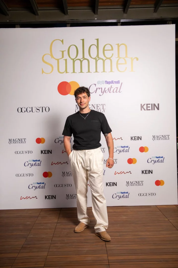 Kıvırcık saçlı genç bir adam, siyah tişörtü, bej pantolonu ve süet makosenleriyle, arka planda büyük altın harflerle yazılmış Golden Summer ve Yapı Kredi Crystal gibi sponsor logolarının bulunduğu şık bir etkinlik alanında kendinden emin bir şekilde poz veriyor.
