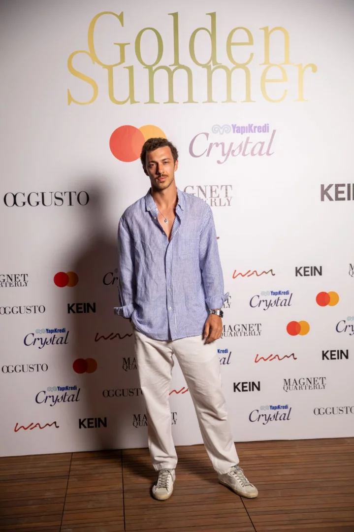 Golden Summer yazısı ve çeşitli sponsor logolarıyla dolu beyaz bir etkinlik fonunun önünde duran, açık mavi çizgili keten gömleği, açık renkli bol pantolonu ve eskitilmiş beyaz spor ayakkabılarıyla şık ve rahat bir görünüm sergileyen genç adam, boynundaki gümüş kolyesiyle kameraya bakıyor.