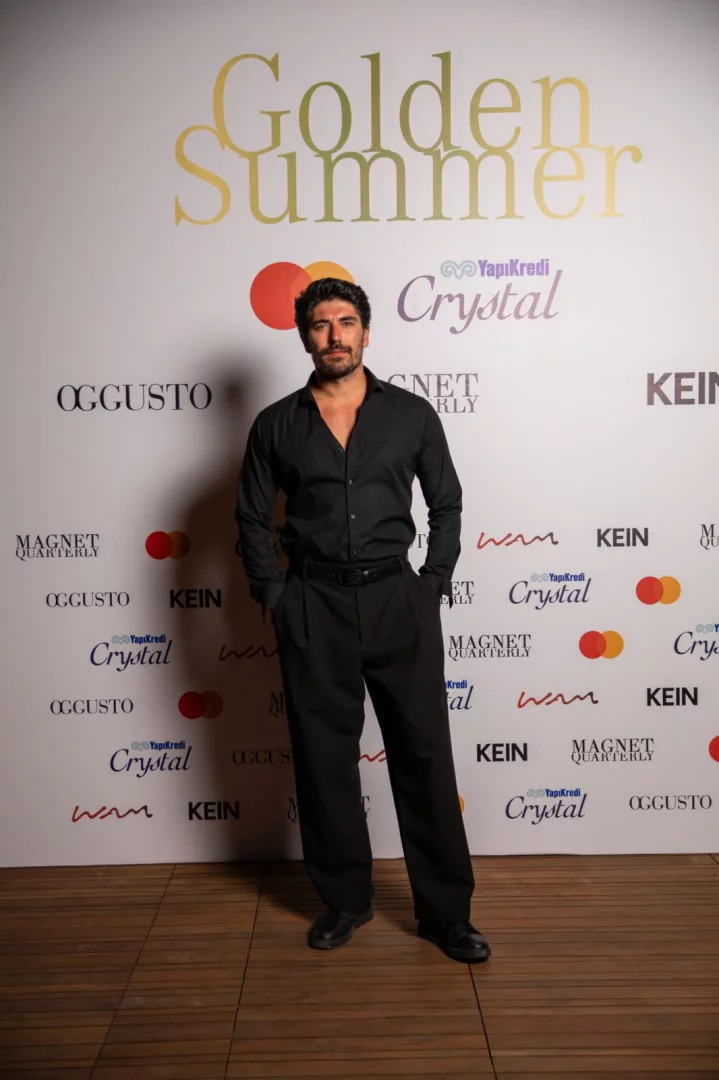 Alp Navruz, siyah gömleği ve pantolonuyla elleri ceplerinde durarak kamera için poz veriyor; arka planındaki beyaz fotoğraf duvarında zarif altın renkli Golden Summer başlığı, OGGUSTO, Magnet Quarterly, Yapı Kredi Crystal ve Mastercard gibi etkinlik ortaklarının logoları dikkat çekiyor.
