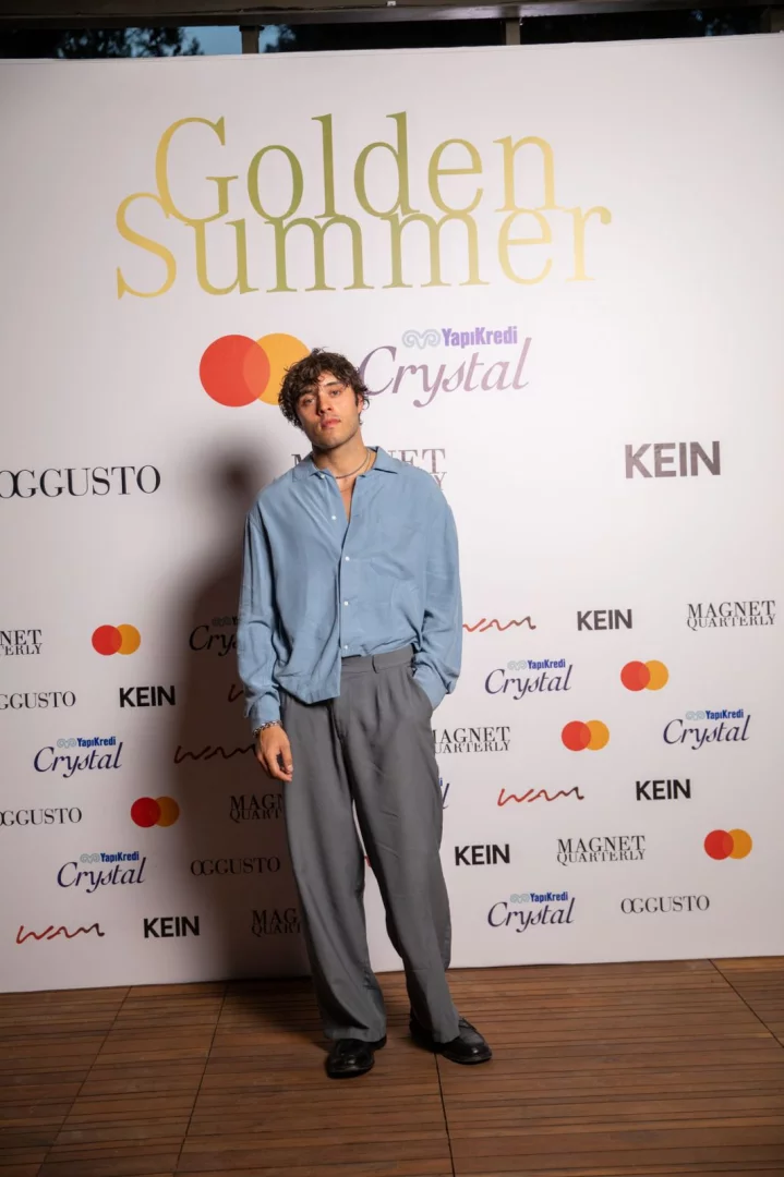 Açık mavi bol bir gömlek ve gri bol bir pantolon giymiş, kıvırcık saçlı genç bir adam, sol elini cebine koymuş halde, YapıKredi Crystal, Mastercard, OGGUSTO ve KEIN gibi sponsor logolarının tekrarlandığı Golden Summer yazılı beyaz bir etkinliğin fonunda kameraya bakıyor.