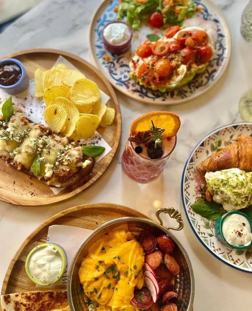 Mermer bir masanın üzerinde, çırpılmış yumurta ve sucuk tavası, kıvırcık patates cipsleriyle servis edilen mavi peynirli tost, cherry domatesli mozarella salatası, kruvasan ve böğürtlenli meyveli içecek gibi birbirinden lezzetli ve çeşitli kahvaltı ürünleriyle dolu zengin bir brunch sofrası sergileniyor.