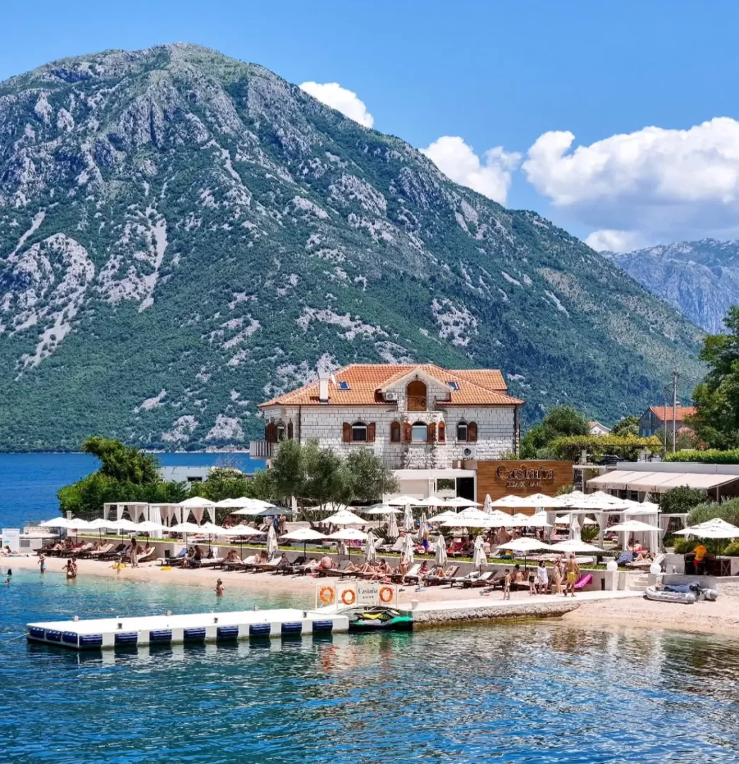 Yemyeşil dağların arasında, berrak mavi bir koyda konumlanan, Castana yazılı otel binasının önündeki plajda çok sayıda beyaz şemsiye, şezlong ve denizde yüzen insanlarla dolu canlı bir tatil beldesi.