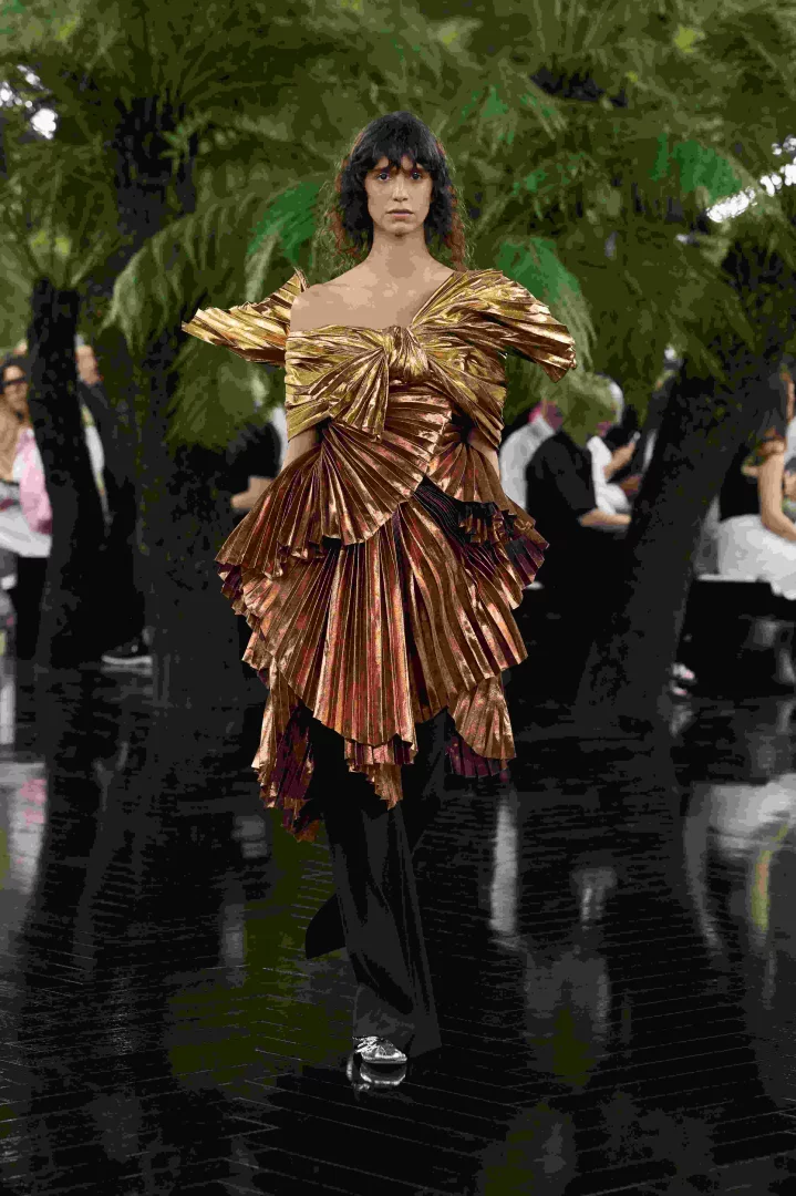 Koyu saçlı bir model, Jonathan Anderson'ın Dior Sonbahar-Kış 2026 Haute Couture koleksiyonundan, Lynda Benglis'in heykellerinden esinlenen, kumaşa heykelsi bir hacim veren drapaj ve pileleme teknikleriyle tasarlanmış, parlak altın-bronz renkte, fiyonk detaylı, çok katmanlı bir bluz ve siyah pantolonla podyumda yürüyor.
