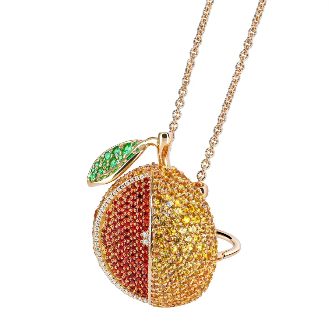 Rose gold zincirde sallanan, dilimlenmiş gibi duran, bir tarafı kırmızı diğer tarafı sarı değerli taşlarla kaplı, ortası pırlanta çizgili gösterişli bir meyve kolye ucu; yeşil yapraklı bu modern tasarım yaz enerjisini yansıtıyor.