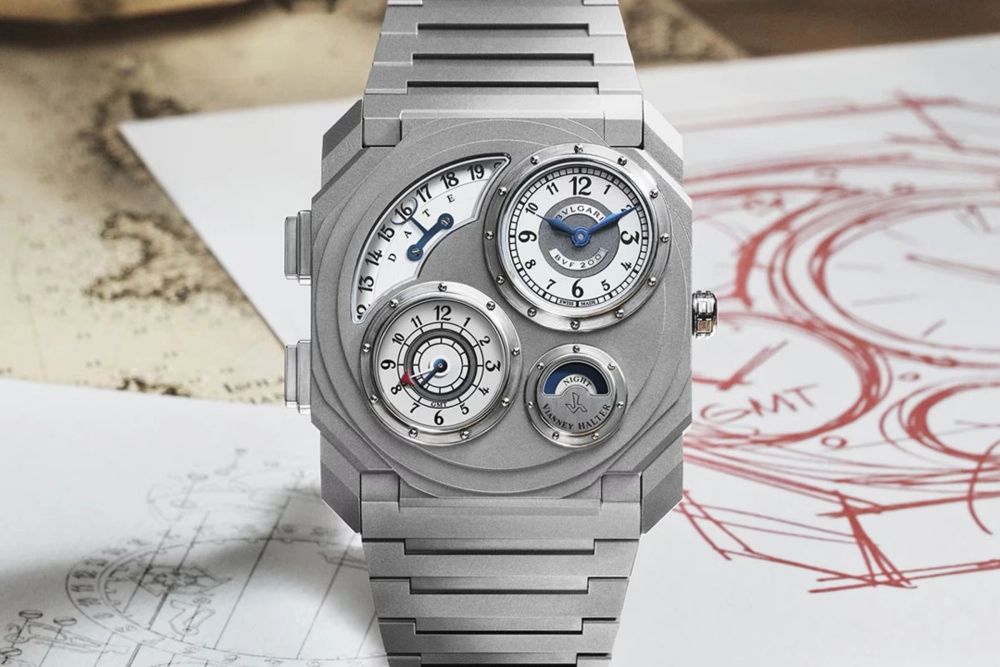 Fırçalanmış gümüş tonlu Bulgari Octo Finissimo Dual Time Vianney Halter kol saati, entegre bileziği ve karmaşık çoklu beyaz alt kadranlarıyla; yerel saat, tarih, GMT ve gece/gündüz göstergelerini detaylandıran tasarımıyla, tasarım çizimlerinin ve haritanın yer aldığı bir zemin üzerinde sergileniyor.