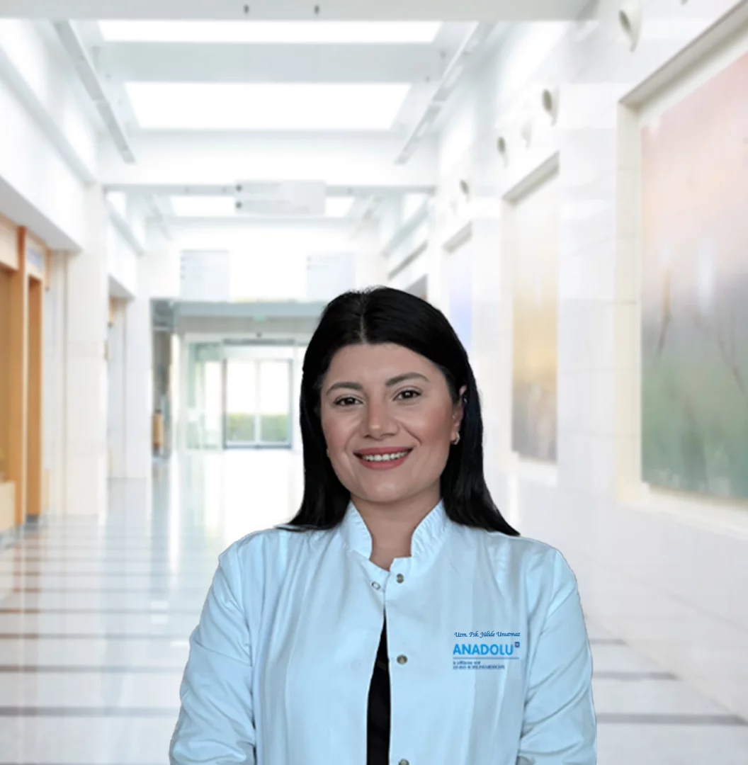 Uzman Psikolog Jülide Unutmaz, modern bir hastane koridorunun aydınlık atmosferinde beyaz önlüğüyle güven veren bir gülümsemeyle durarak, Tükenmişlik Sendromu ve İş Yeri Deformasyonu hakkında makalede okuyuculara rehberlik ediyor.