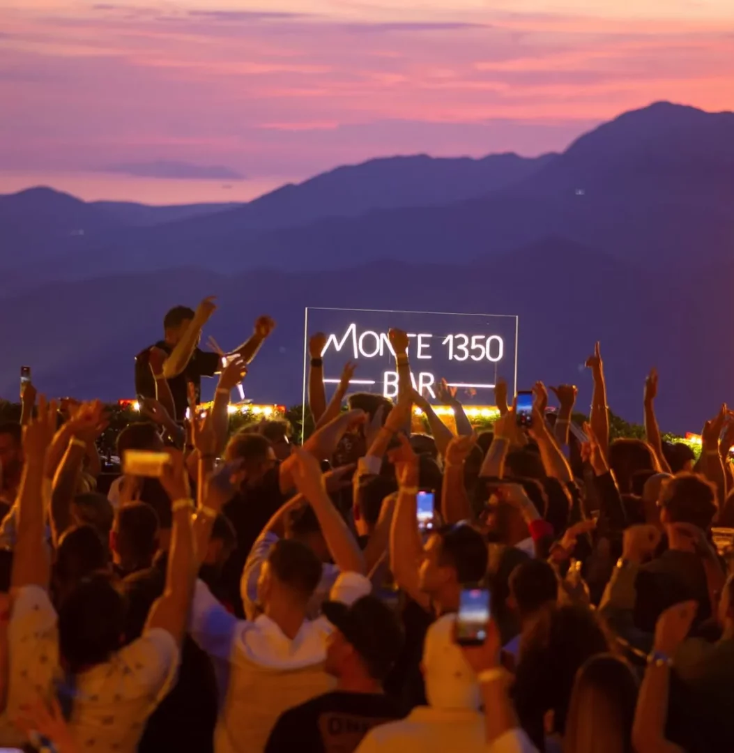 Gün batımının pembe ve mor tonlarındaki gökyüzünün altında, dağ manzaralı bir açık hava bar etkinliğinde, DJ'in önünde Monte 1350 Bar yazan neon tabelanın ışığında ellerini havaya kaldırmış coşkulu bir kalabalık dans ediyor ve eğleniyor.