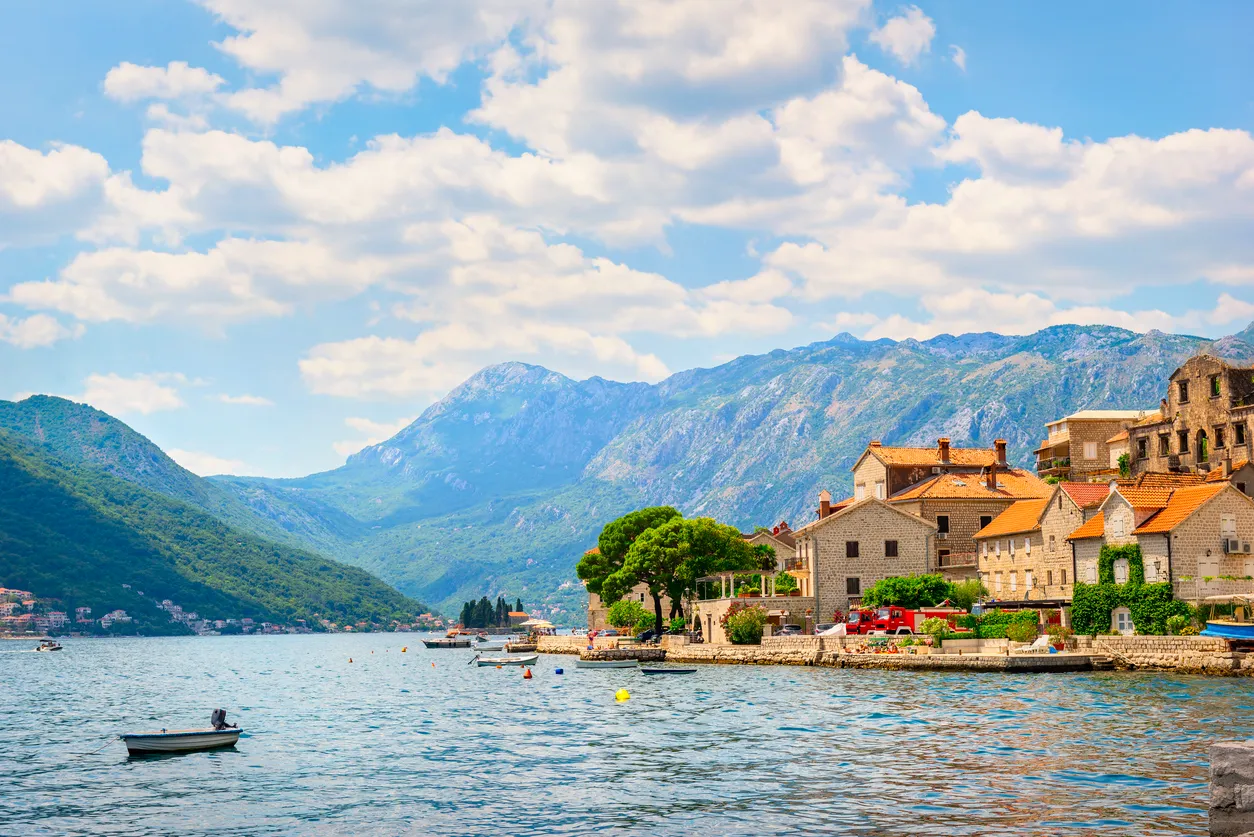 Perast kasabasının kırmızı çatılı tarihi taş evleri, berrak turkuaz körfez suları boyunca uzanırken, yemyeşil dağlar ve gökyüzündeki beyaz bulutlar büyüleyici bir Akdeniz manzarası oluşturuyor.