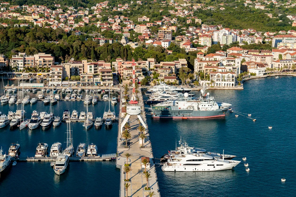 Porto Montenegro marinasının kuşbakışı görünümünde, masmavi denizde sıralanmış lüks yatlar ve yelkenliler, palmiye ağaçlarıyla süslü uzun bir iskele ile arka plandaki yeşil tepelere yayılan çatılı binalar göz alıcı bir şekilde birleşiyor.