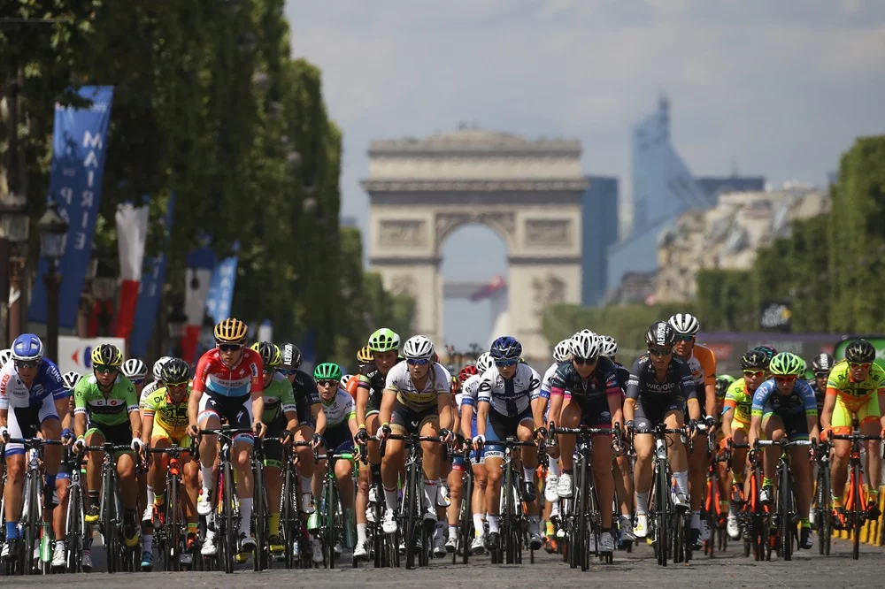 Paris'in ikonik Zafer Takı'nın net bir şekilde göründüğü Champs-Élysées üzerinde, çeşitli renklerdeki formalarıyla büyük bir bisikletçi grubu, güneşli bir günde Tour de France'ın final etabında toplu halde hızla pedal çeviriyor.