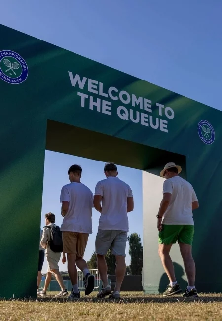 Güneşli bir günde, WELCOME TO THE QUEUE yazılı yeşil bir kemerin altından geçerek Wimbledon turnuvasının köklü geleneklerinden biri olan sıraya doğru ilerleyen birkaç kişi görülüyor, bu sıra gerçek Wimbledon deneyimini yaşamak isteyenler için turnuvanın ruhunu paylaşmanın bir parçası.