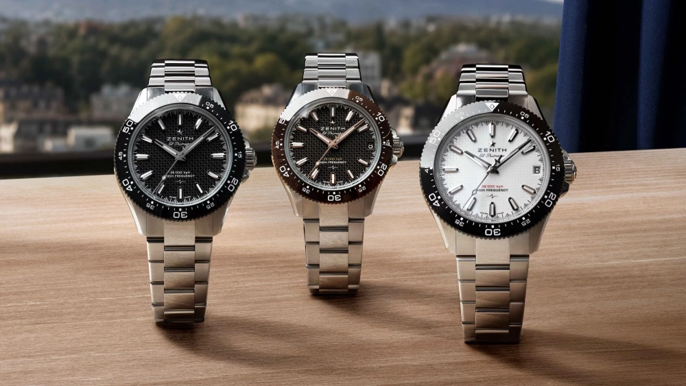 Ahşap bir yüzey üzerinde sergilenen, biri siyah, biri kahverengi ve biri beyaz kadrana sahip üç farklı Zenith Chronomaster Sport kol saati, parlak çelik bilezikleri ve uyumlu renklerdeki bezelleriyle dikkat çekiyor, arka planda ise bulanık bir şehir manzarası ve koyu mavi bir perde görünüyor.