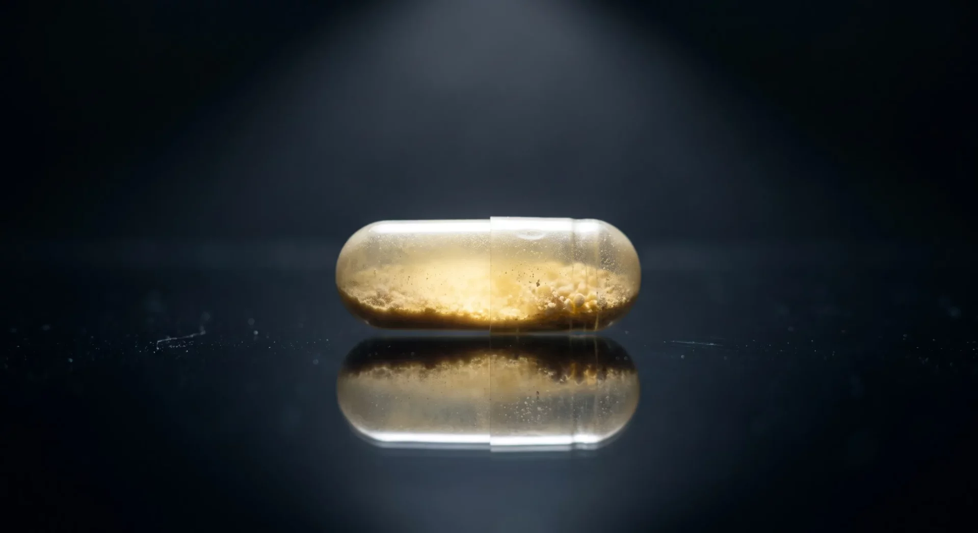 Bir spot ışığıyla yukarıdan aydınlatılmış, içinde sarımsı kahverengi granül tozlar bulunan şeffaf bir B6 vitamini kapsülü, yansıtıcı koyu renkli bir yüzeyde yatay olarak duruyor ve yüzeydeki toz partikülleri ışıkla vurgulanıyor.